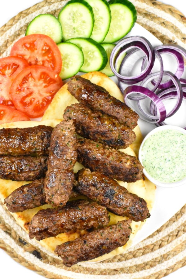 Platter of seekh kebab with onion, tomato, cucumber, pita and raita.
